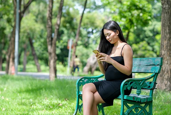 young pregnant woman sits on park bench looking at free open adoption app on her phone